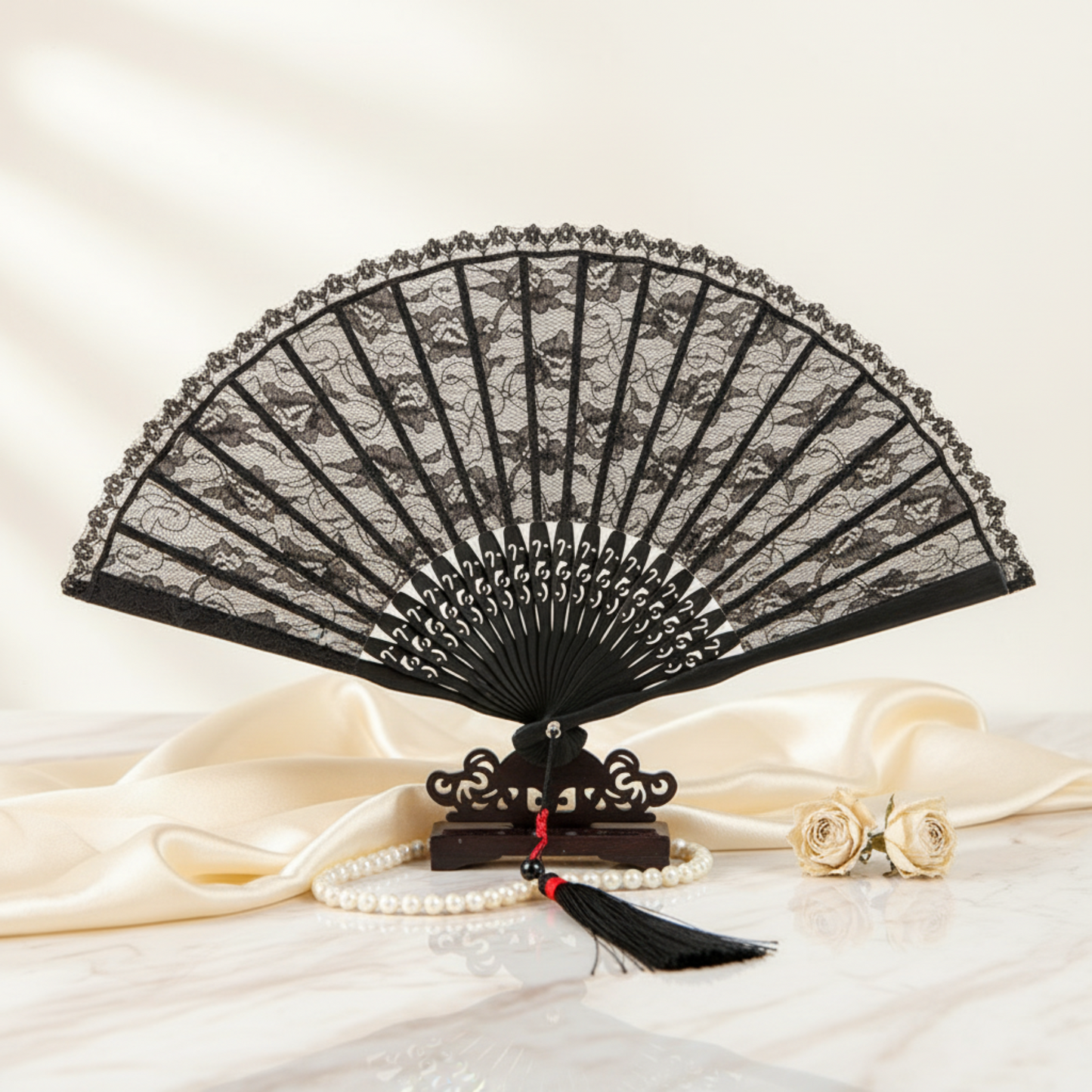 A black lace folding hand fan displayed open on a small wooden stand, with cream satin fabric, a pearl strand, and two gold roses in the foreground against a soft cream background.