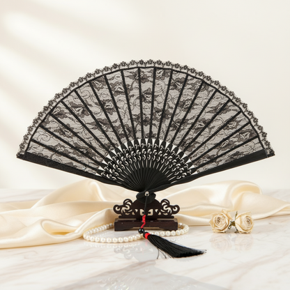 A black lace folding hand fan displayed open on a small wooden stand, with cream satin fabric, a pearl strand, and two gold roses in the foreground against a soft cream background.